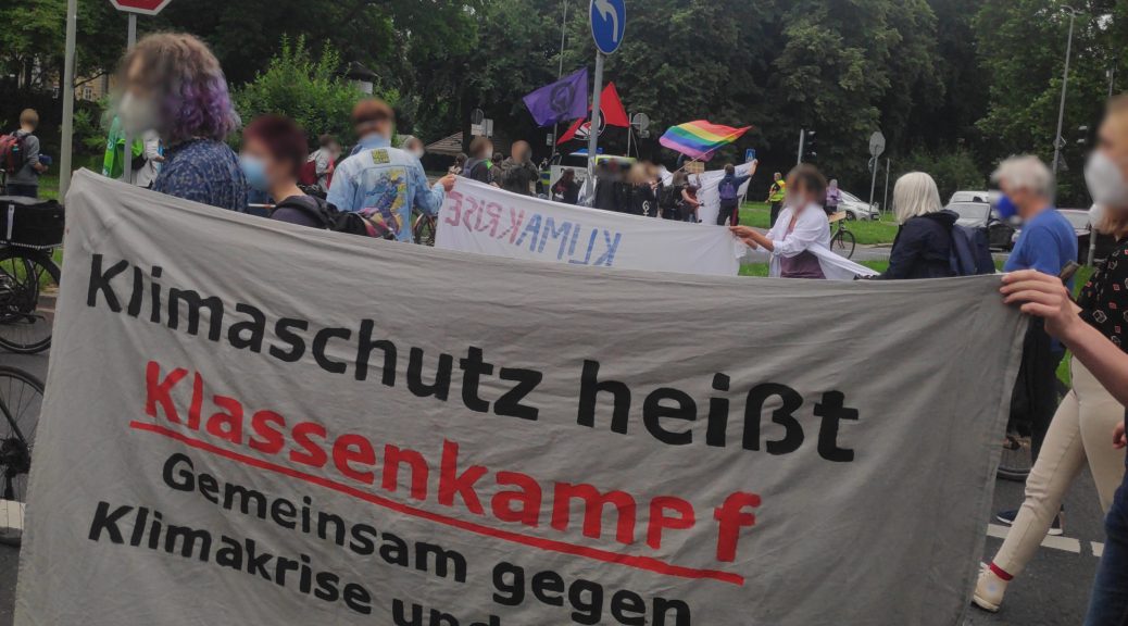 Demo Foto, zeigt großes Transparent mit dem Text: "Klimaschutz heißt Klassenkampf". Im Hintergrund ist eine Prideflag, eine Antifa-Fahne und eine Lila Fahne mit Feminismus-Faust